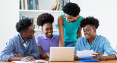 A teacher looks over a group of smiling students working on a computer